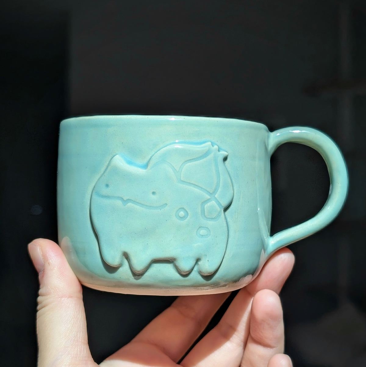 A light blue-green ceramic mug with a raised bulbasaur design, held in a person's hand against a dark background.